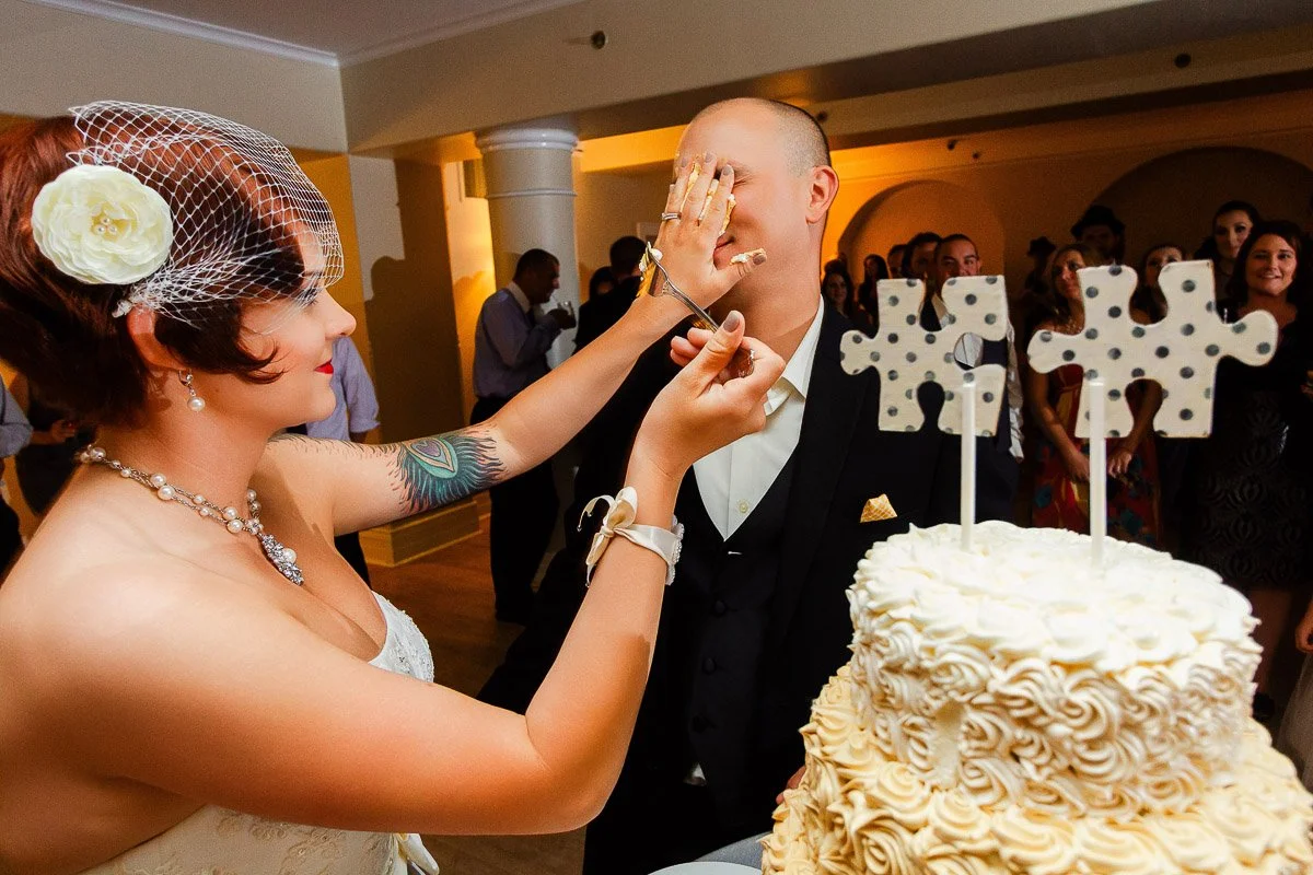 A bride playfully smears cake on the groom's face at a wedding reception. A decorated cake with puzzle piece toppers is in the foreground, guests in the background laugh.