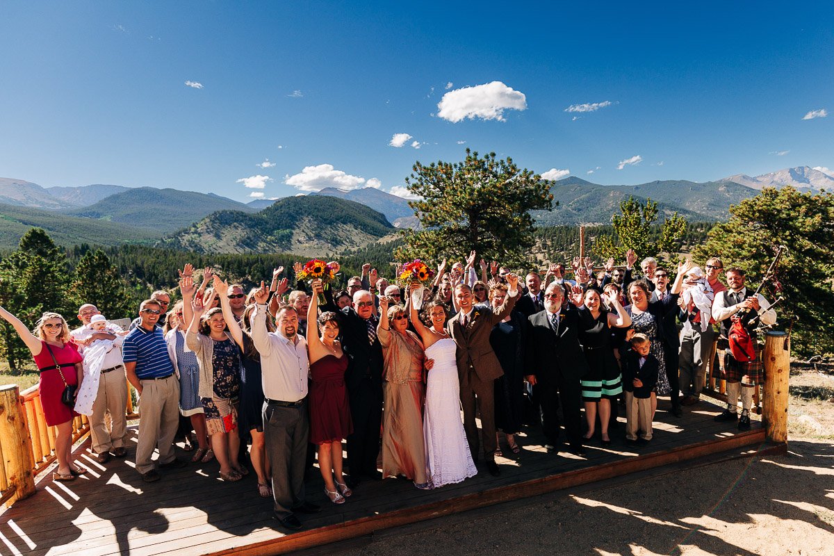 A joyful wedding party poses outdoors on a sunny day with mountains in the background. Guests cheer with raised hands, celebrating the couple in the center.