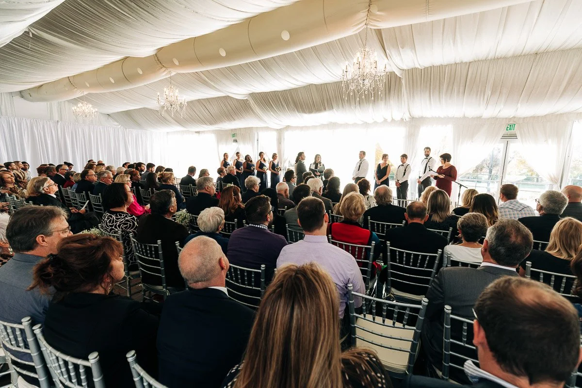 A large audience sits in rows facing a group of people standing at the front under a white draped ceiling with chandeliers, creating an elegant, formal atmosphere.