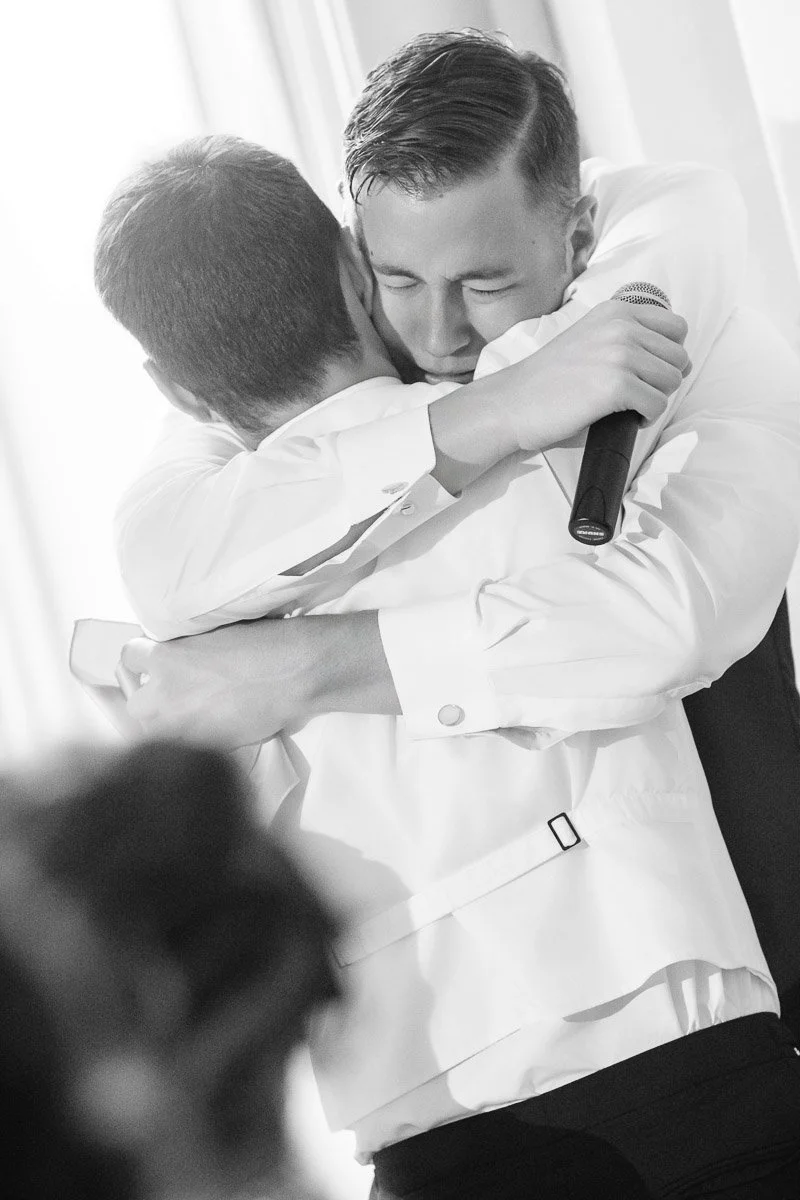 Two men in white shirts embrace tightly, one holding a microphone. Their expressions convey deep emotion and joy. The scene is intimate and heartfelt.