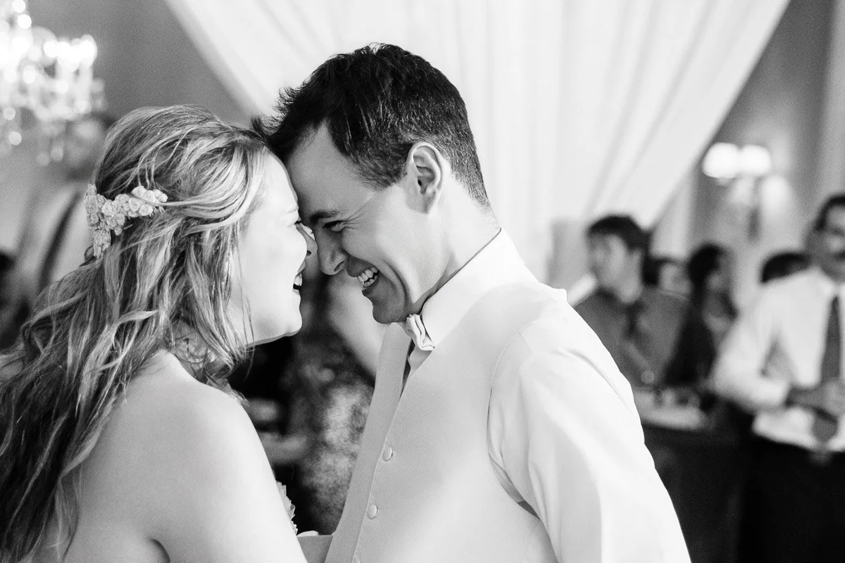 A joyful couple is captured sharing a close, affectionate moment while dancing at their wedding. The bride has long hair with floral accents. Guests are blurred in the background.