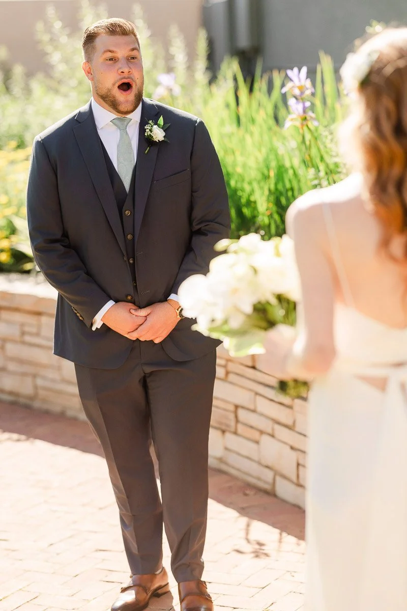 A bride and groom sit outdoors, the groom in a suit is playfully covered by the bouquet held by the bride. She is wearing a white dress and birdcage veil, making a playful expression.