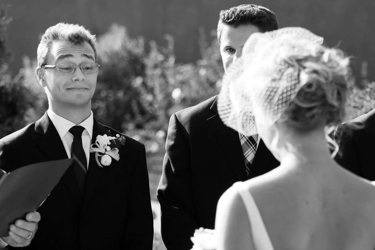 A groom with a playful smirk faces a bride in a veil during an outdoor wedding ceremony, with a best man smiling behind in the bright sunlight.