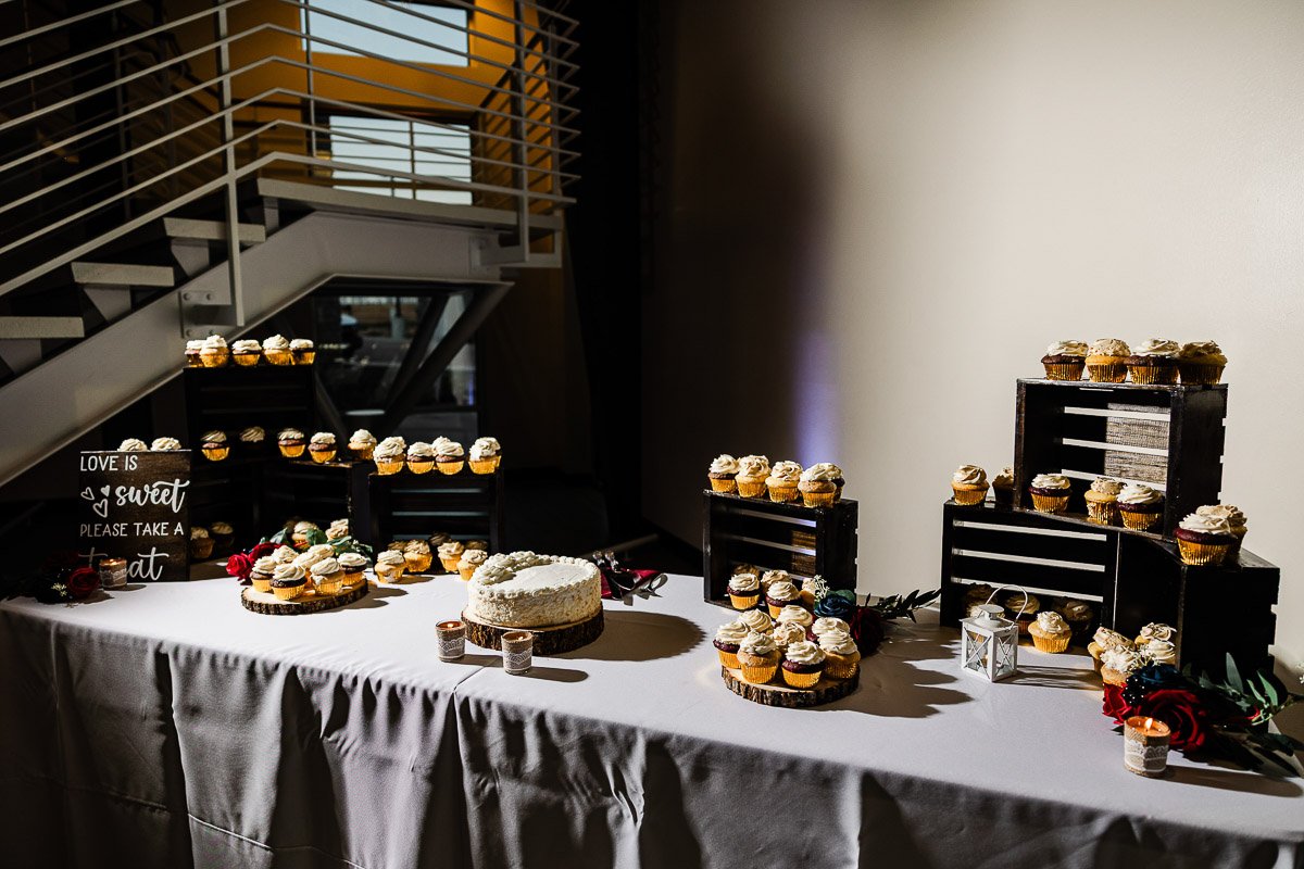 A dessert table with assorted cupcakes on wooden stands and a white cake on a pedestal. A sign reads, "Love is sweet, please take a treat." Warm, inviting ambiance.