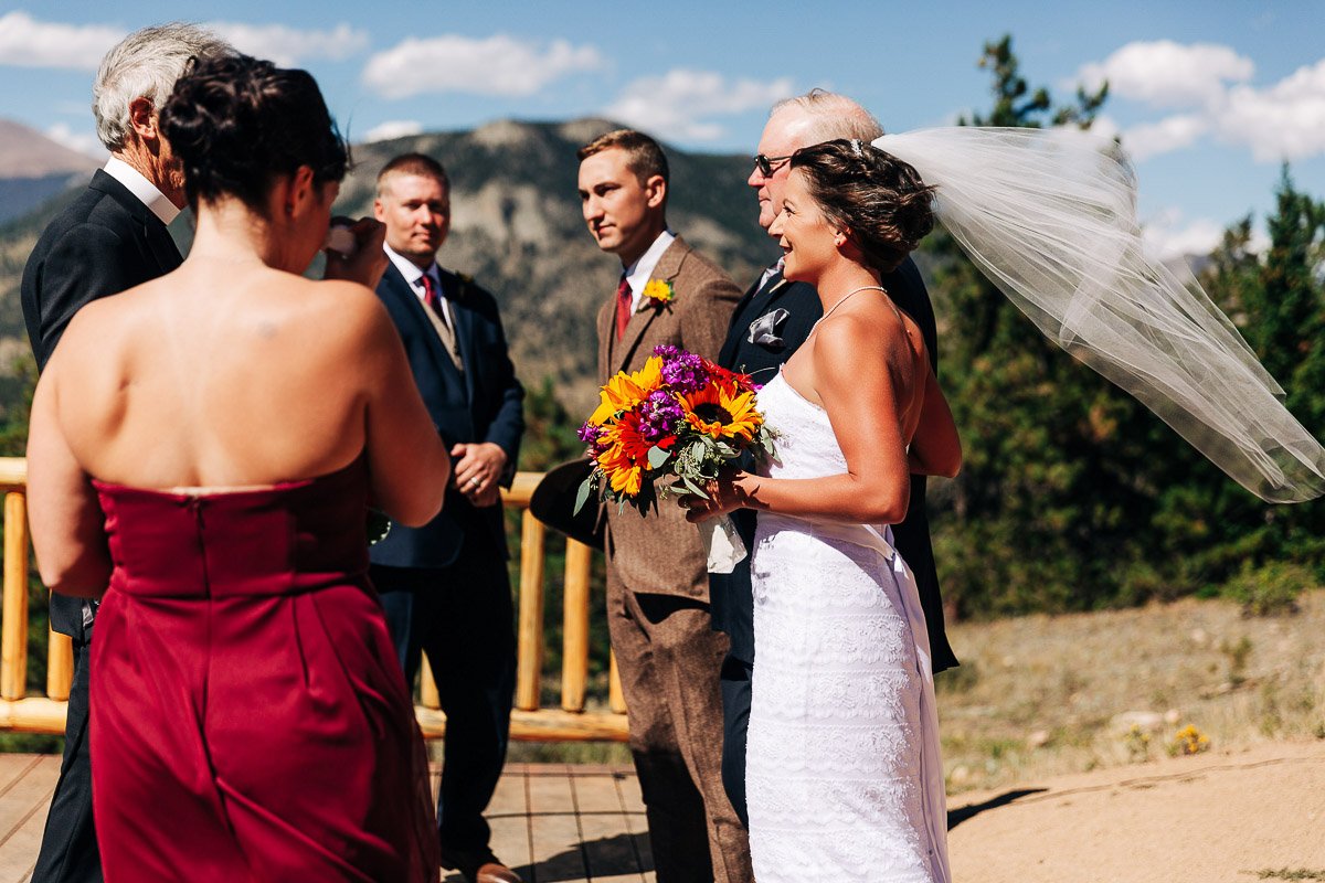 A bride in a white dress with a flowing veil holds colorful flowers, standing beside a groom in a brown suit. They are on a scenic mountain overlook, accompanied by a few guests. The mood is joyful and celebratory.