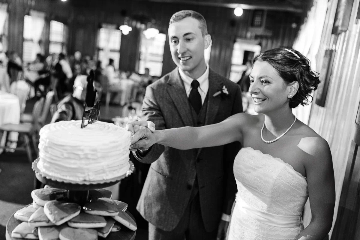 A bride and groom happily cut a tiered wedding cake together. The bride wears a strapless dress and pearl necklace, creating a joyful wedding ambiance.