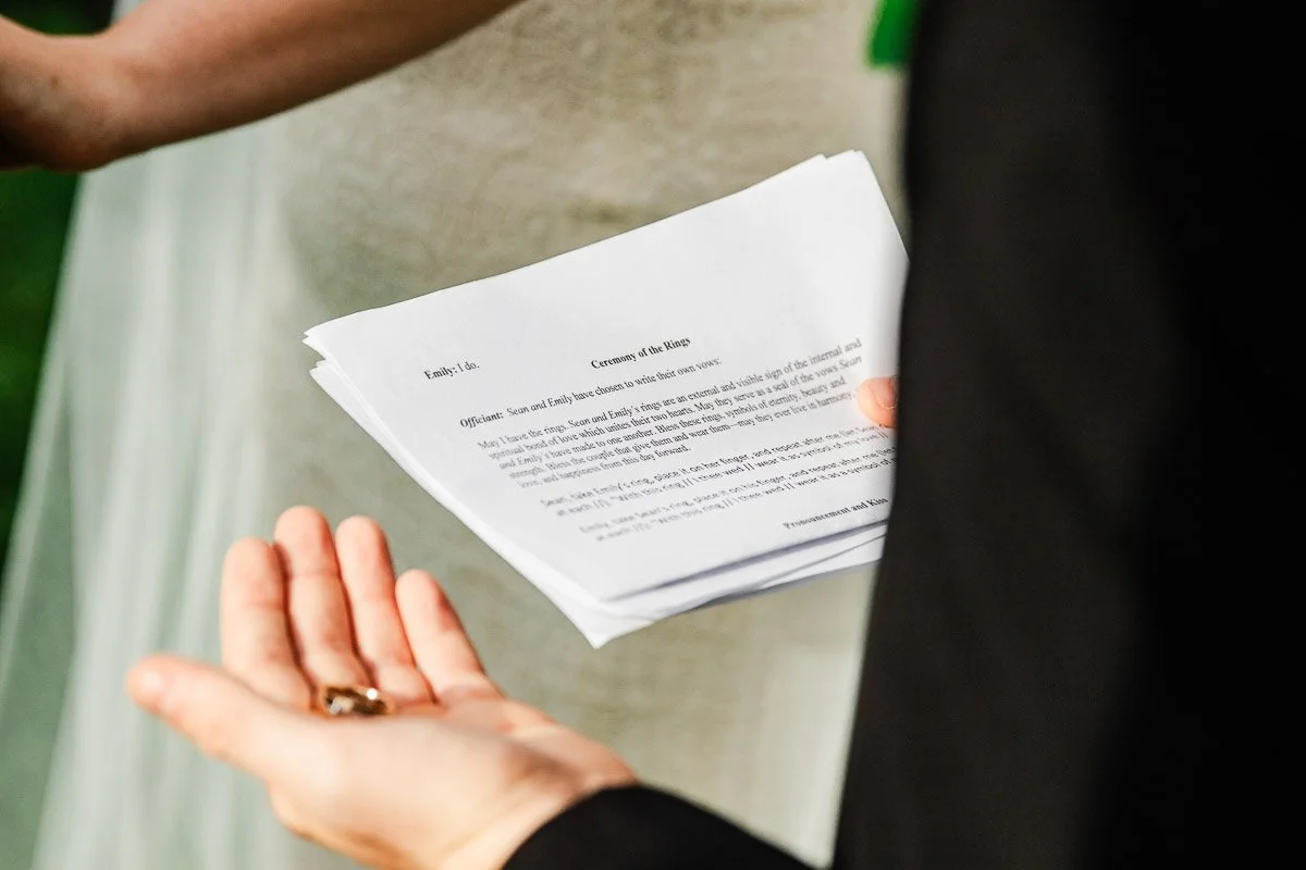 Close-up of a wedding ceremony. A person in a black suit holds a script titled "Ceremony of the Rings," with another hand holding wedding rings.