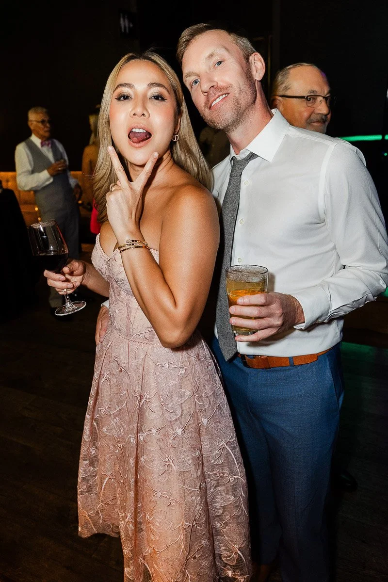 A woman in a pink lace dress playfully poses with a man in a white shirt and blue pants, both holding drinks at a lively social event.