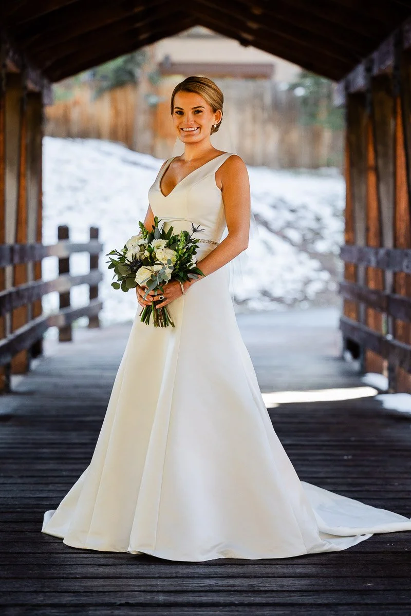A bride in a white gown smiles while holding a bouquet of white and green flowers. She stands on a rustic wooden bridge with a snowy background.