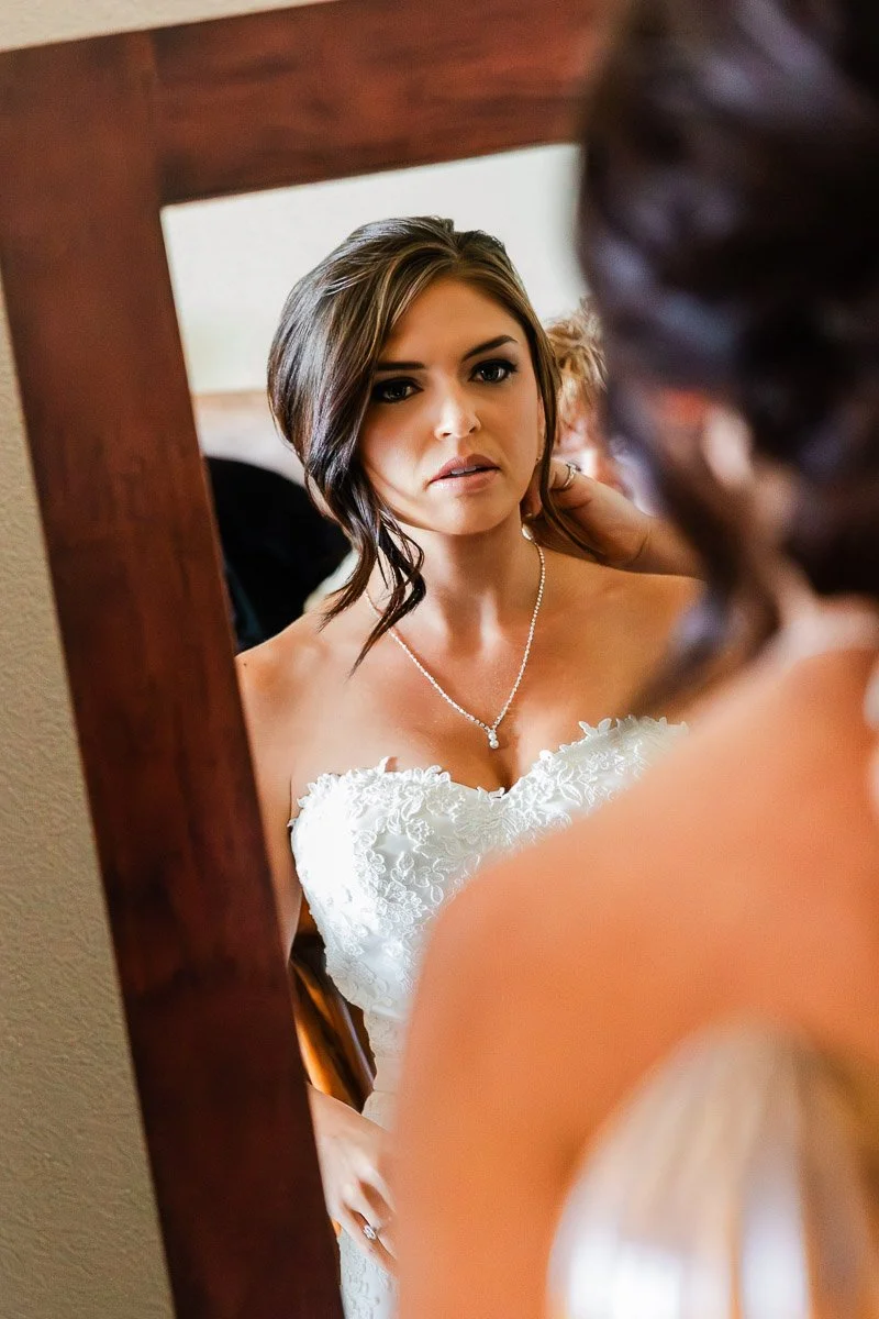 A bride in a strapless lace gown gazes into a wooden-framed mirror, her expression thoughtful. Her hair is styled elegantly, and a necklace adorns her neck.