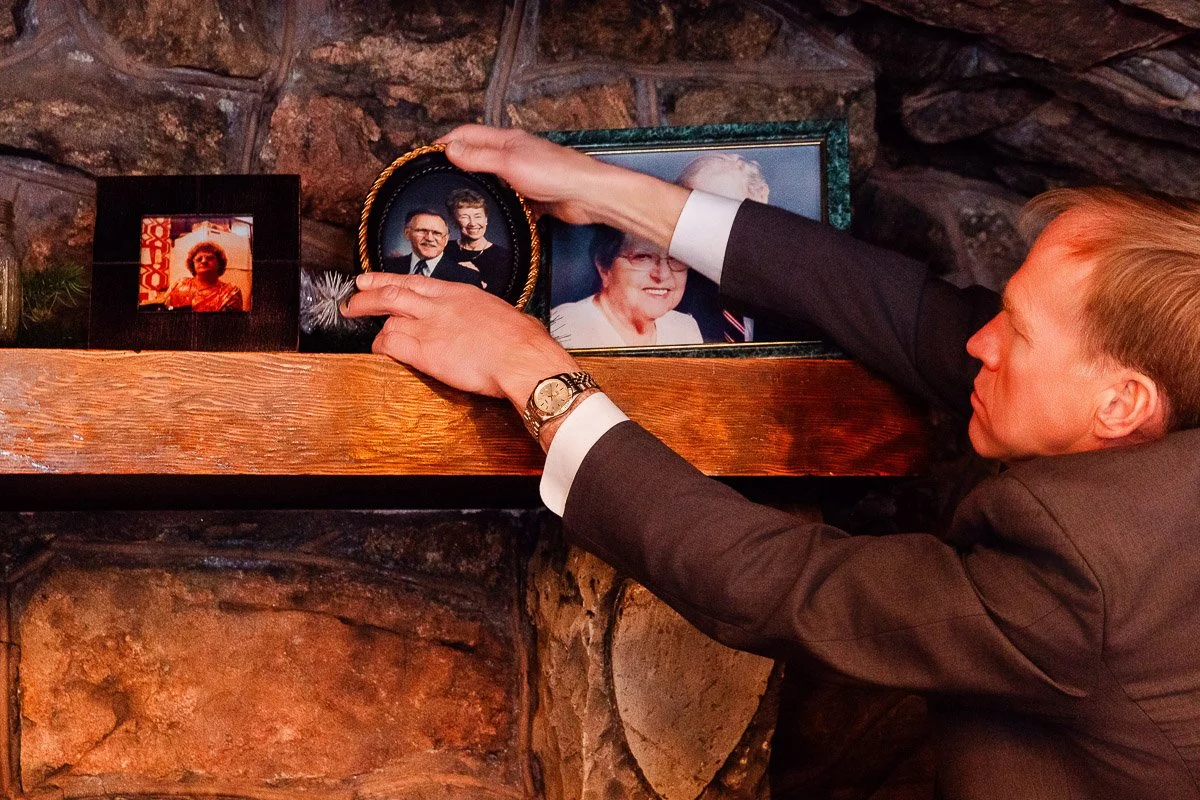A man wearing a suit carefully arranges family photos on a wooden mantel. The setting suggests a warm, nostalgic atmosphere with cherished memories.