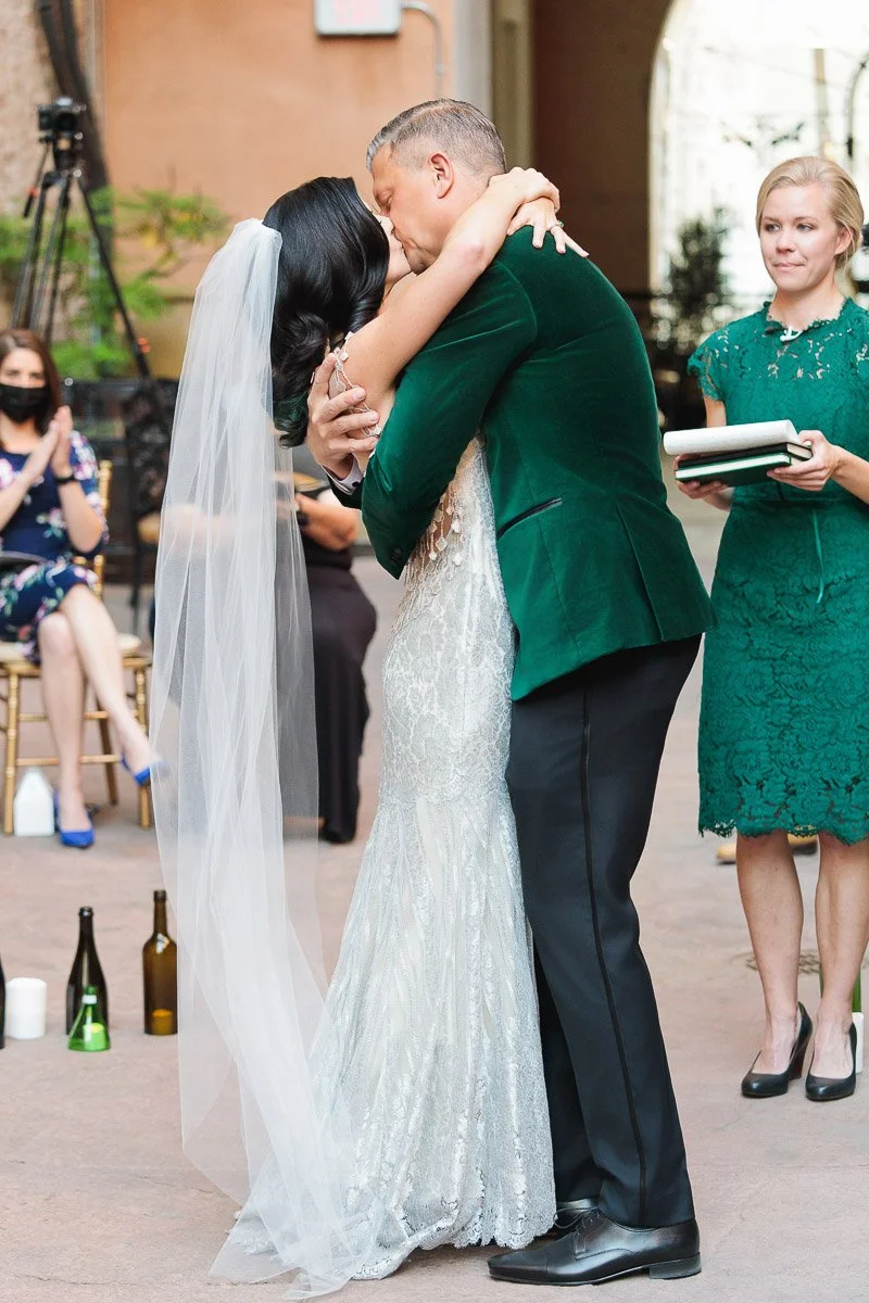 A couple embraces tenderly; the bride in a lace gown and veil, the groom in a green velvet jacket. Onlookers watch with happiness.