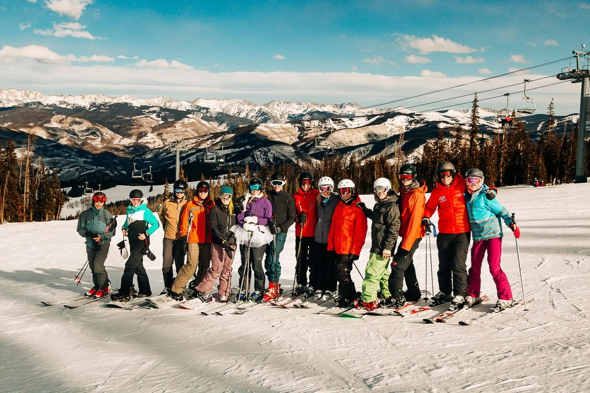 Group of skiers in colorful gear standing on a snowy mountain slope. Chairlift and pine trees in background, with mountains under a blue sky.