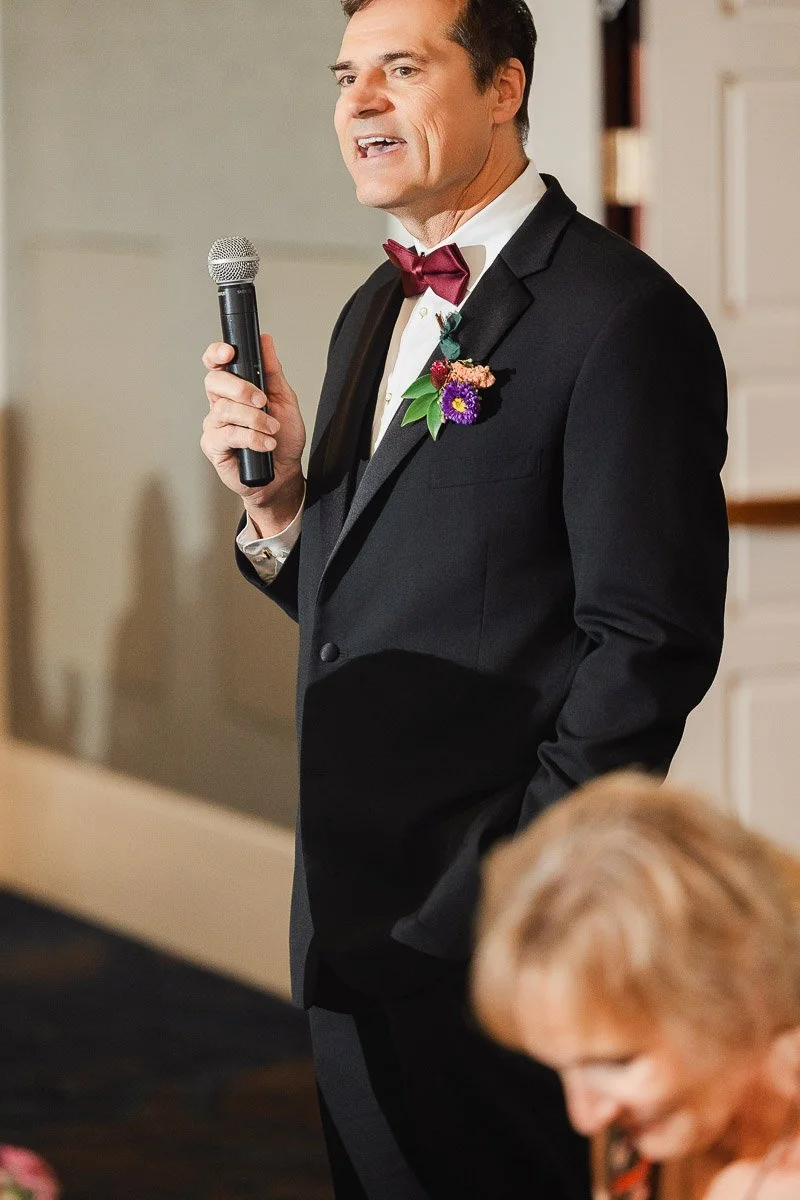 Man in a tuxedo and bow tie holding a microphone, delivering a speech. A boutonniere decorates his lapel. A woman in focus is in the foreground.