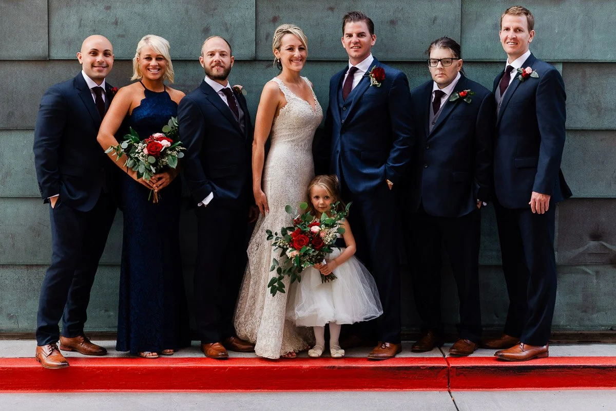 A bride and groom stand smiling with their wedding party in elegant attire. The flower girl holds a bouquet, adding charm to the joyful group scene during a Coohills Restaurant wedding in Denver, Colorado.