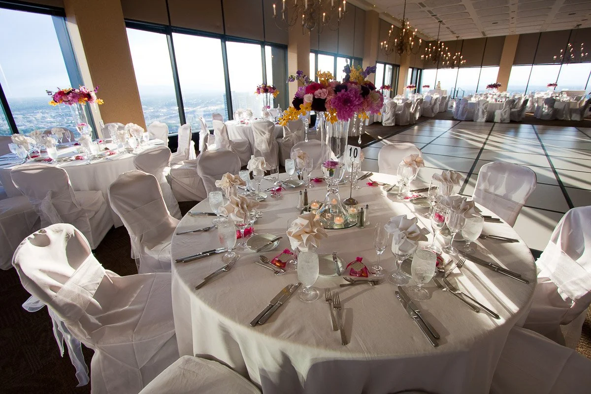 Elegant banquet hall with round tables adorned with white linens and vibrant floral centerpieces. Sunlight streams through large windows, creating a warm, inviting ambiance.