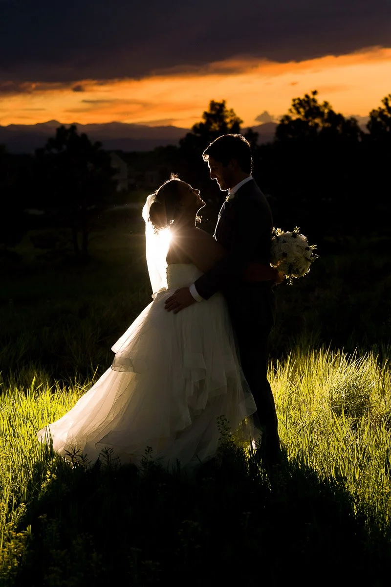 A bride and groom stand close in a field during sunset, creating a romantic silhouette. The bride's veil and dress glow in the warm light during a Villa Parker wedding in Parker, Colorado