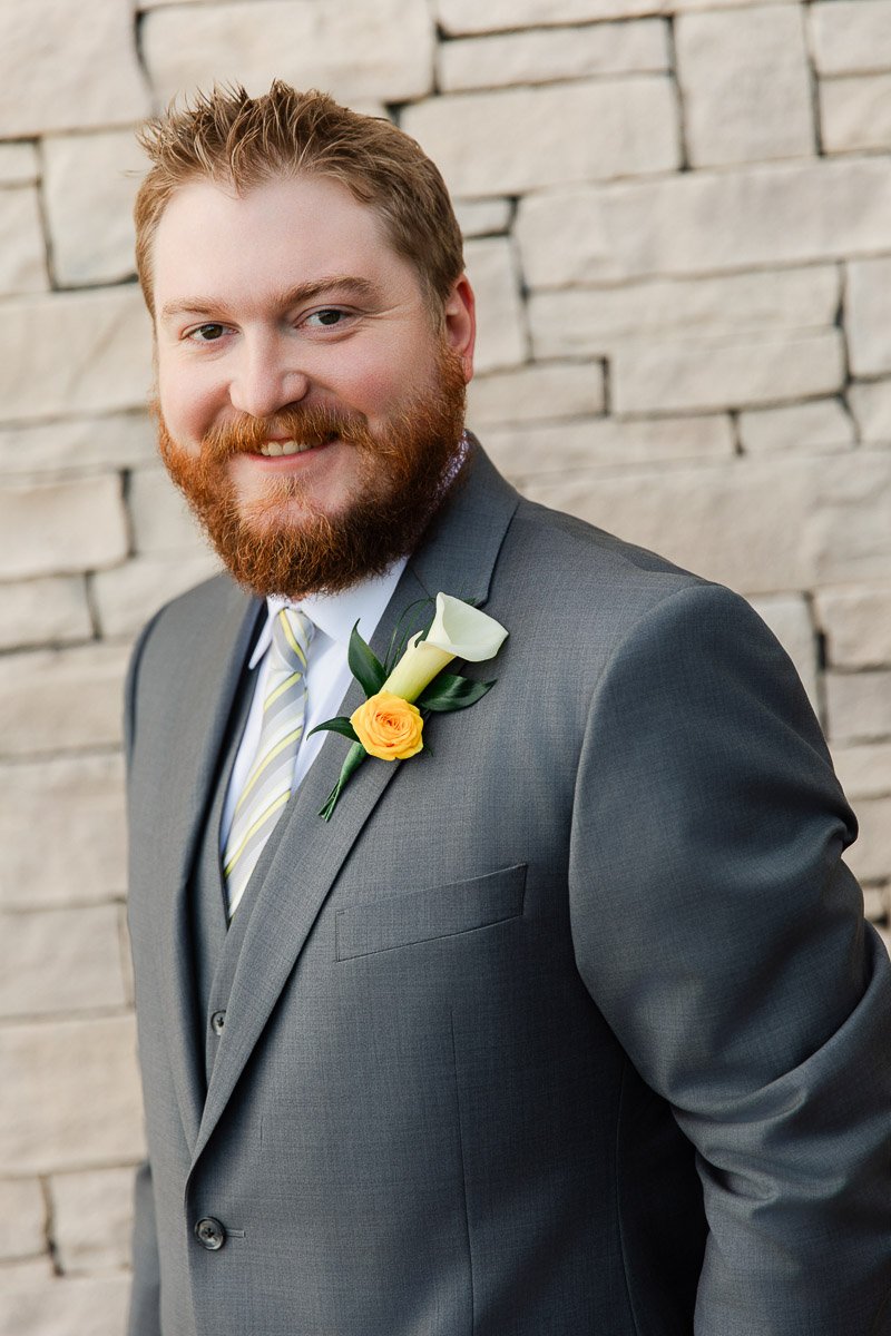 A man with a red beard smiles warmly while wearing a grey suit with a yellow and white flower boutonniere, standing against a stone wall.