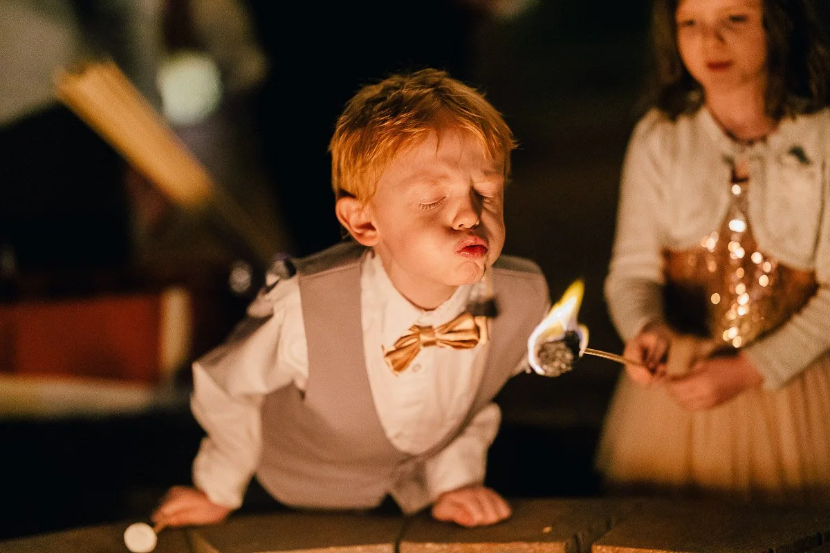 A young boy with red hair, wearing a bow tie and vest, blows on a flaming marshmallow. A girl in a sparkly dress watches with amusement. Night setting, warm light.