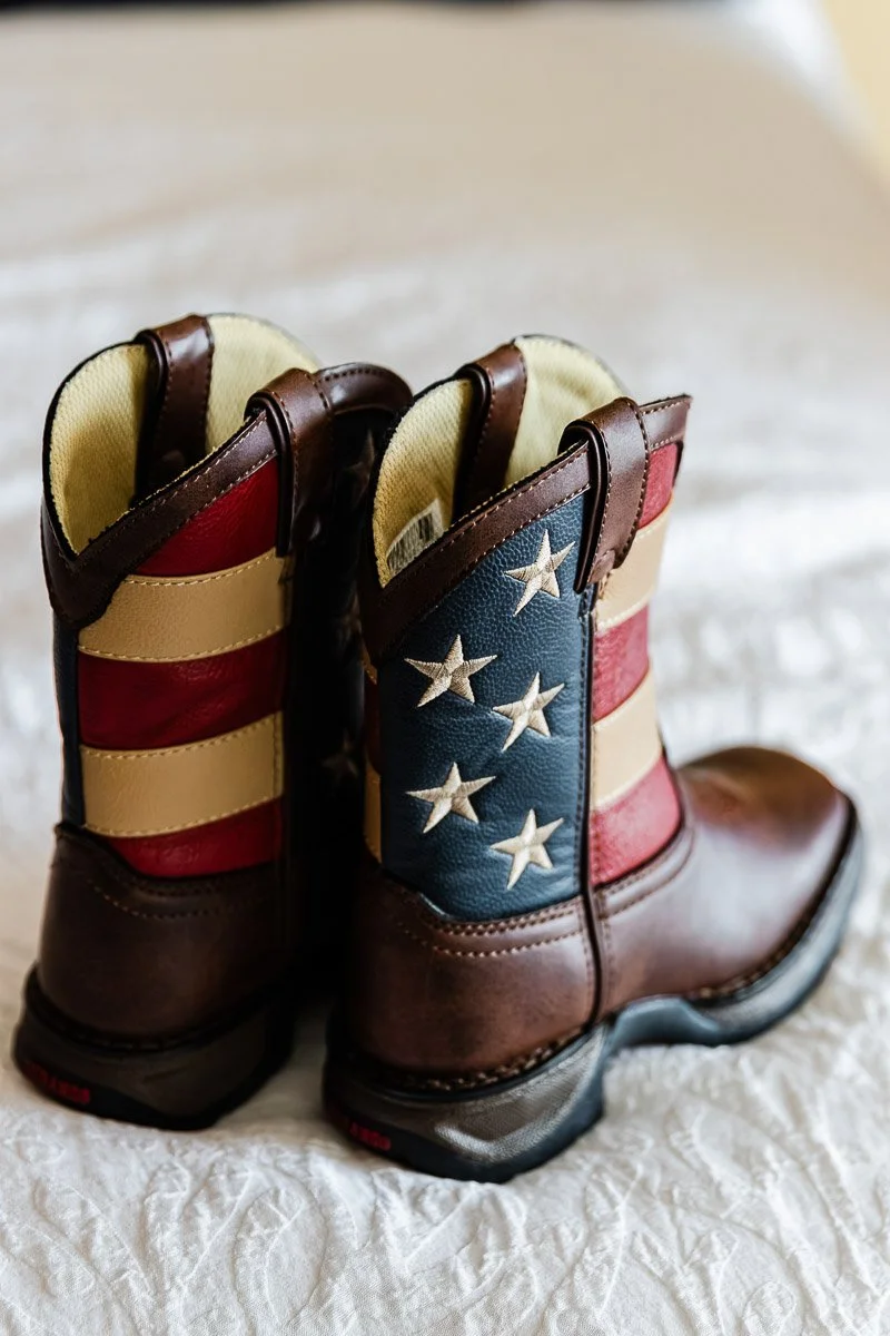 Children's cowboy boots with American flag design, featuring red, white, and blue stripes and stars, resting on a textured white bedspread. Patriotic theme.