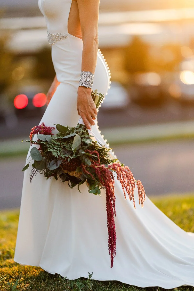 A bride in a fitted white gown with a jeweled bracelet holds a lush bouquet. The sunset casts a warm glow, creating a romantic and serene atmosphere.
