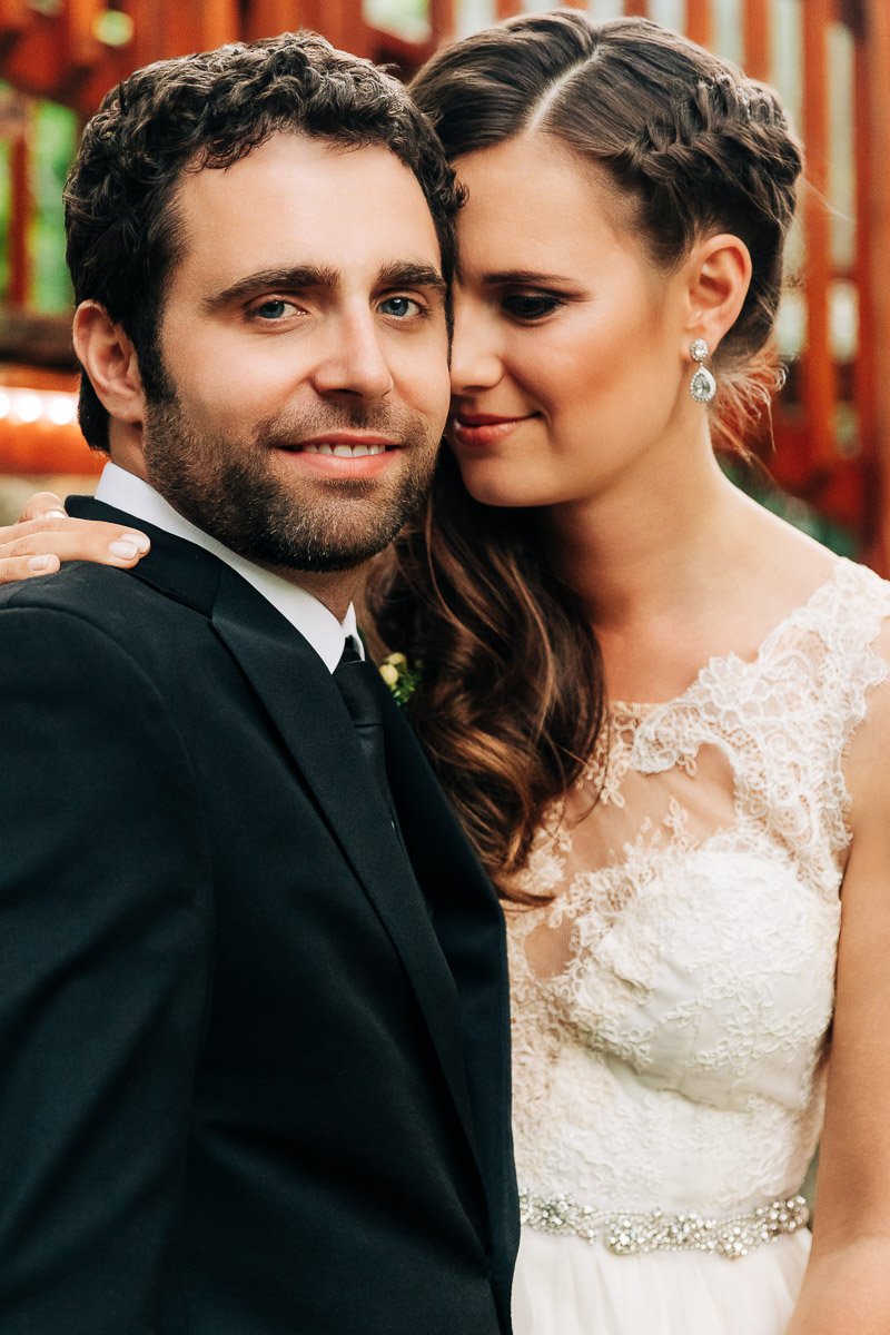 A smiling groom in a black suit embraces his bride in a lace gown with braided hair. The outdoor setting has a warm, joyful ambiance.