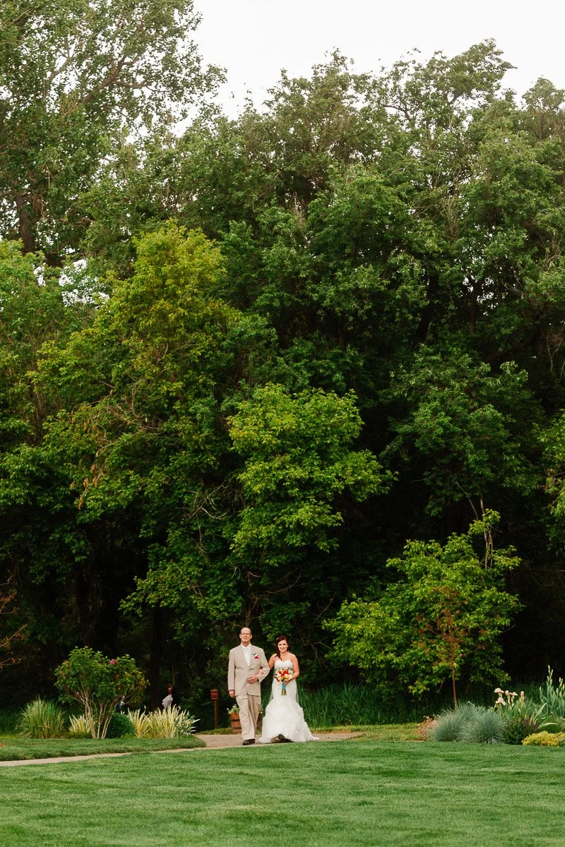 A bride and her father walk together on a garden path, surrounded by lush green trees. The bride holds a bouquet, conveying a joyful, serene atmosphere.