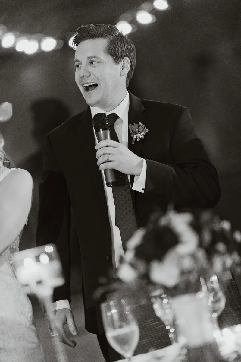 A man in a suit joyfully speaks into a microphone at a wedding. He's smiling, with blurred lights in the background, creating a festive atmosphere.