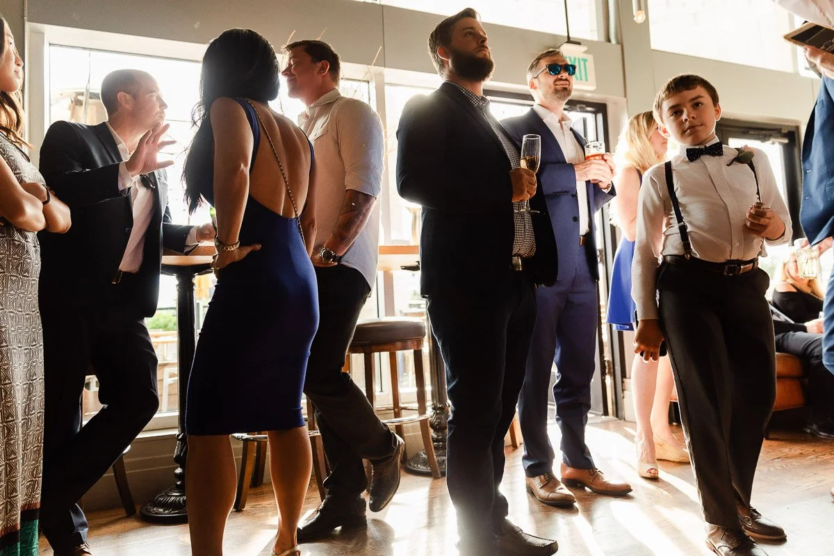 A lively gathering in a sunlit room shows people dressed formally. A young boy in suspenders walks confidently, while others engage in animated conversation.