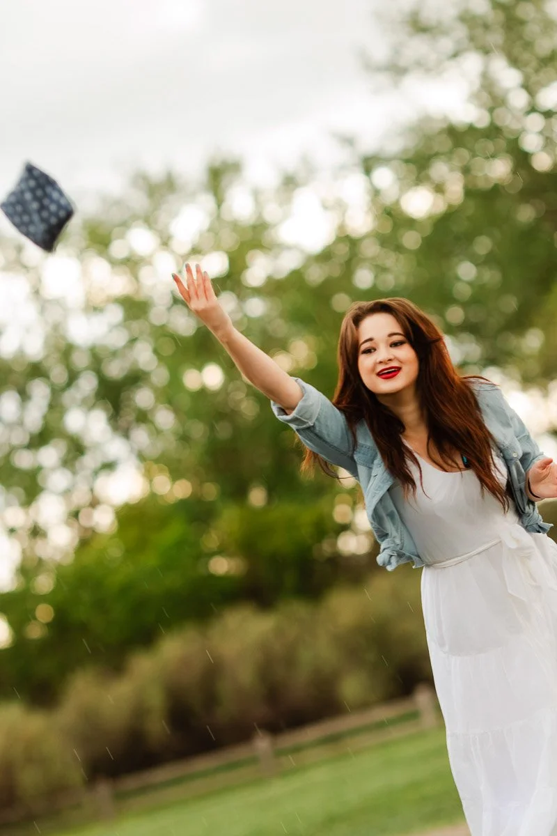 A woman joyfully tosses a small purse in the air in a park. She wears a white dress and denim jacket, with a bright smile. The background features lush green trees.