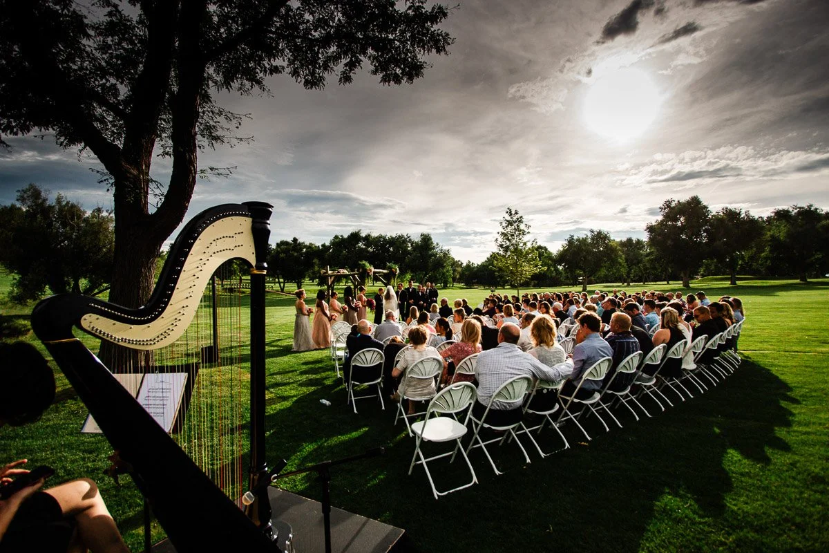 Outdoor Lakewood Country Club wedding ceremony on a sunny day. Guests sit in rows of white chairs on green grass, with a harp in the foreground. Trees and clouds in the background.