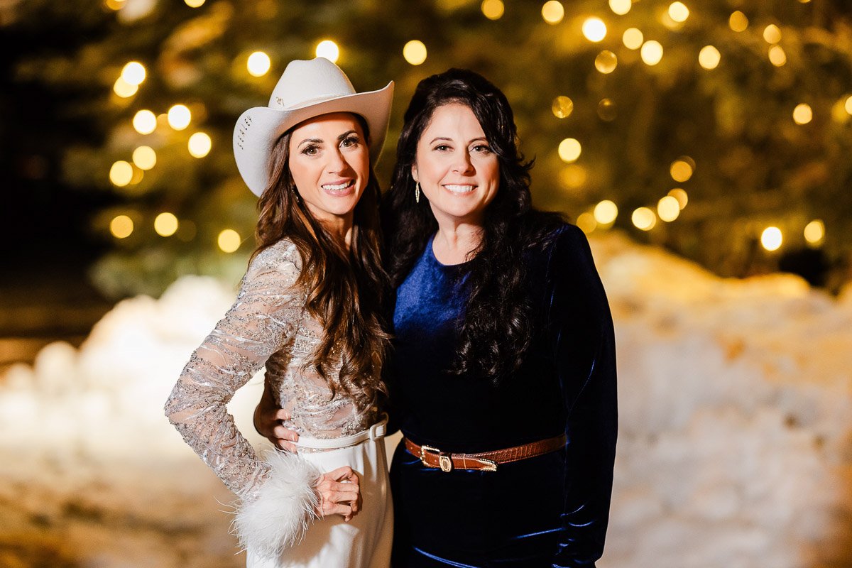 Two women stand smiling at night, surrounded by softly lit bokeh lights. One wears a white cowboy hat and sparkly outfit, the other a dark blue dress.