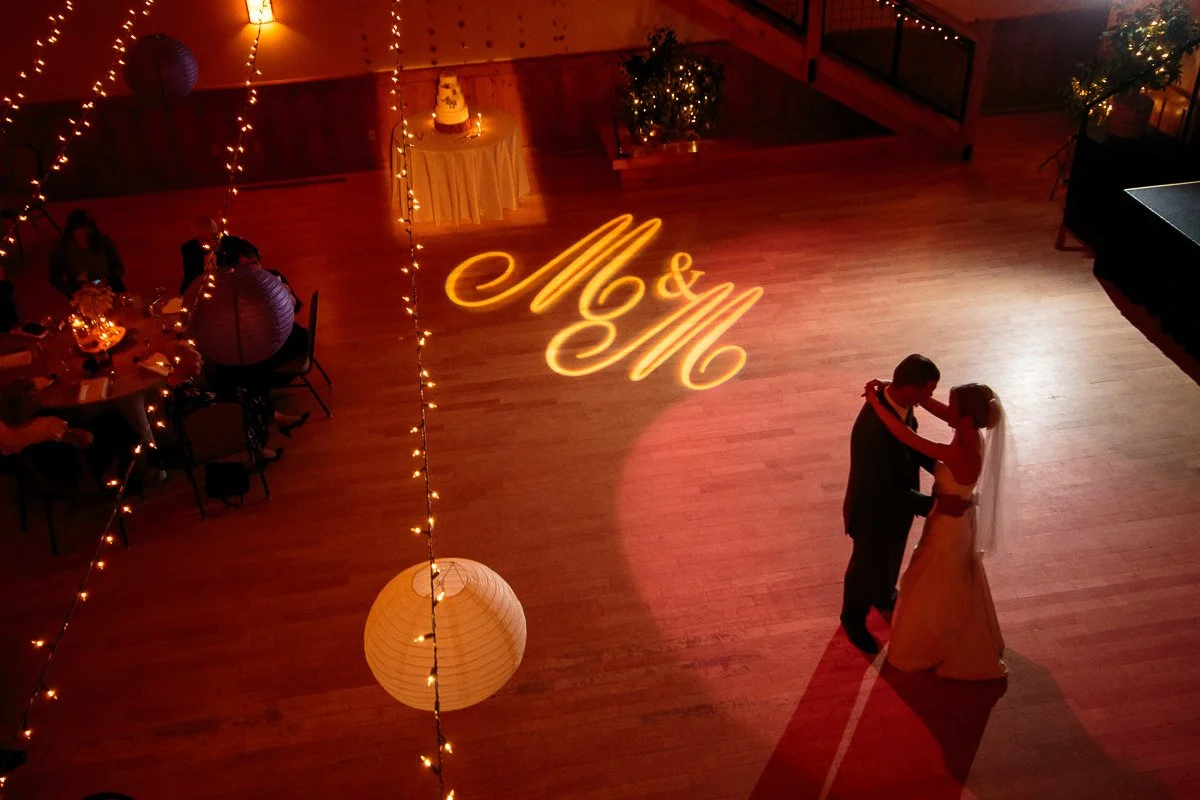 A bride and groom share a first dance on a warmly lit ballroom floor. Initials "M&M" are projected in elegant script. Guests sit around softly illuminated tables.
