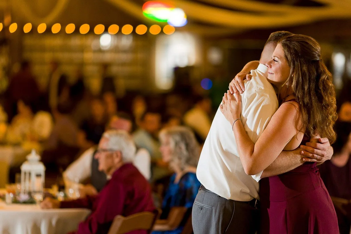 A couple embraces warmly on a dance floor, surrounded by softly lit, blurry background of guests seated at tables. The scene conveys joy and celebration.