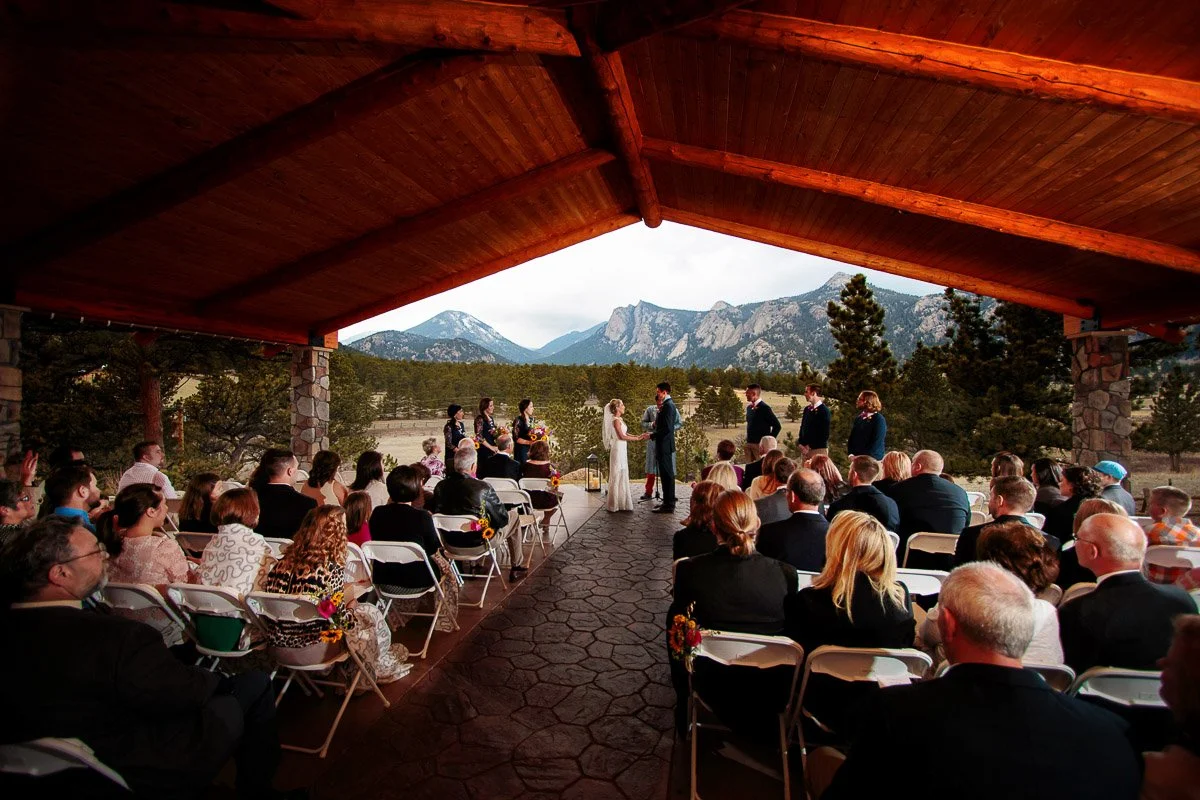 Black Canyon Inn wedding ceremony under a wooden pavilion overlooking Lumpy Ridge mountains. A couple stands at the altar surrounded by guests seated on white chairs, creating an intimate, rustic setting.