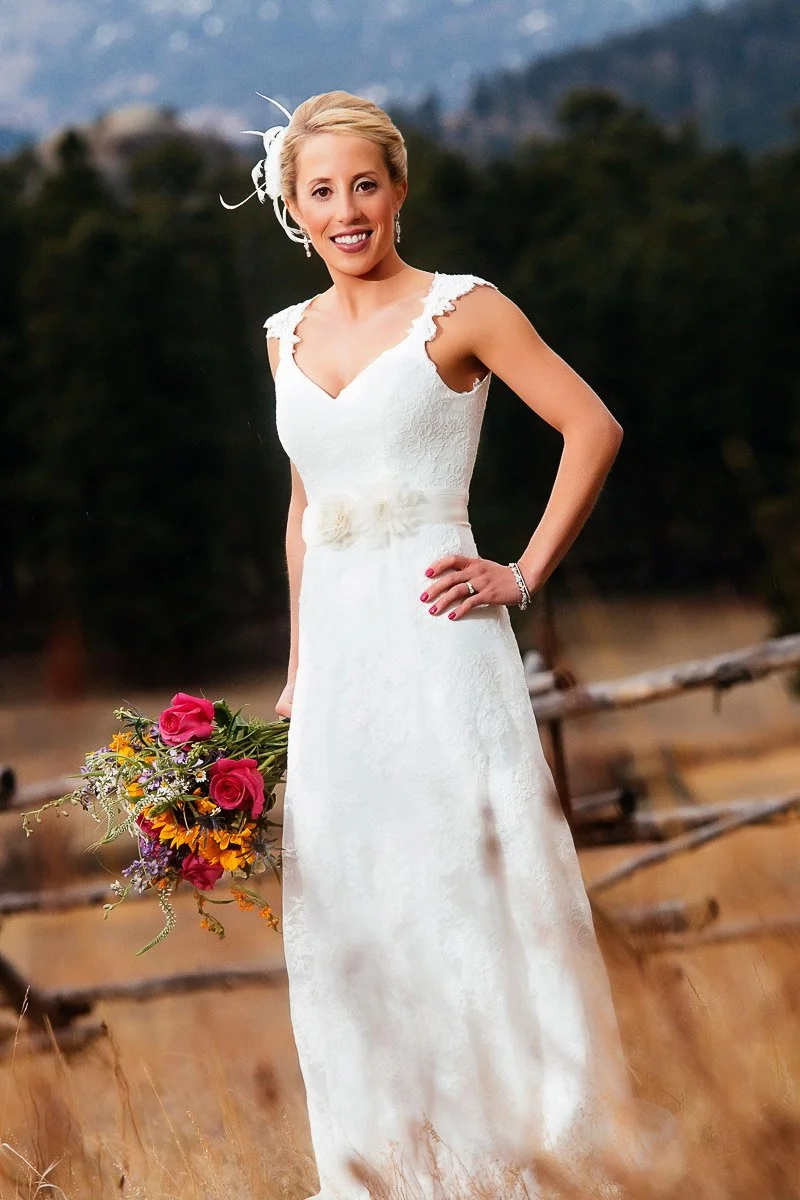 Bride in a white lace dress holding a colorful bouquet, standing in a field with a blurred wooden fence and forest in the background. She smiles warmly.