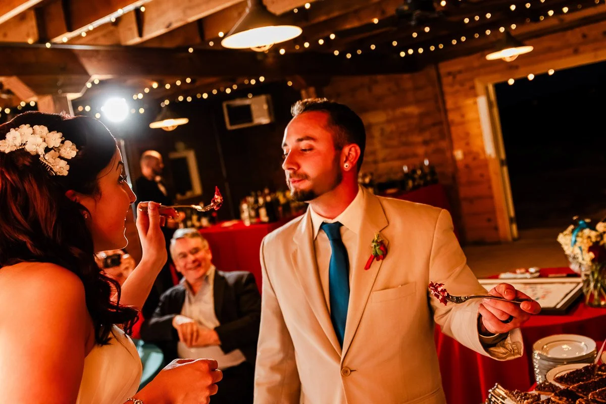 A bride and groom sharing cake in a warmly lit, rustic setting. The bride holds a fork playfully, while the groom leans in, smiling. A guest watches and laughs.