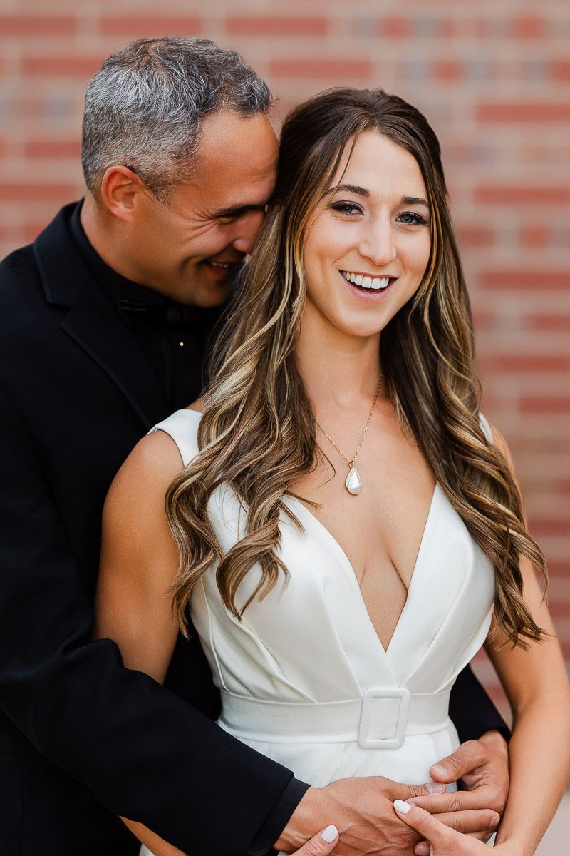 A smiling woman in a white dress stands in front of a smiling man in a black suit, embracing her from behind. They are in front of a brick wall.
