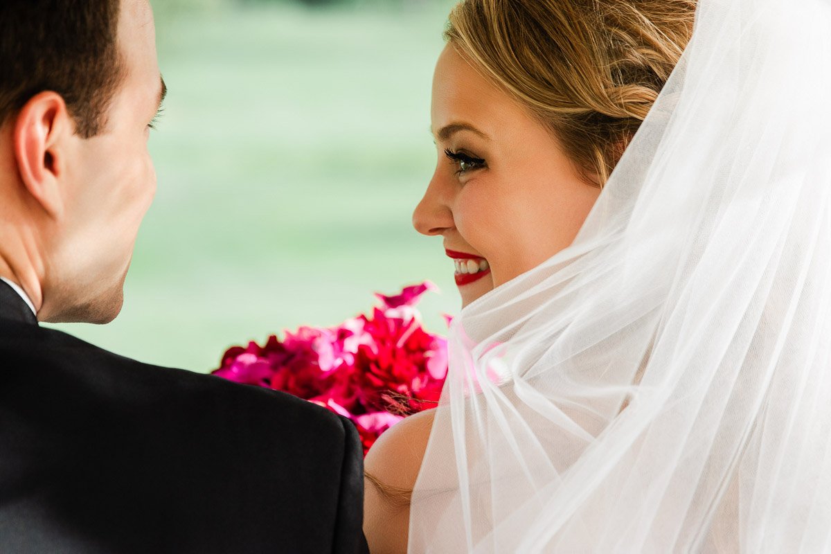 Bride smiling at groom, wearing a white veil, holding red flowers. Bright, joyful atmosphere with a soft, green background.