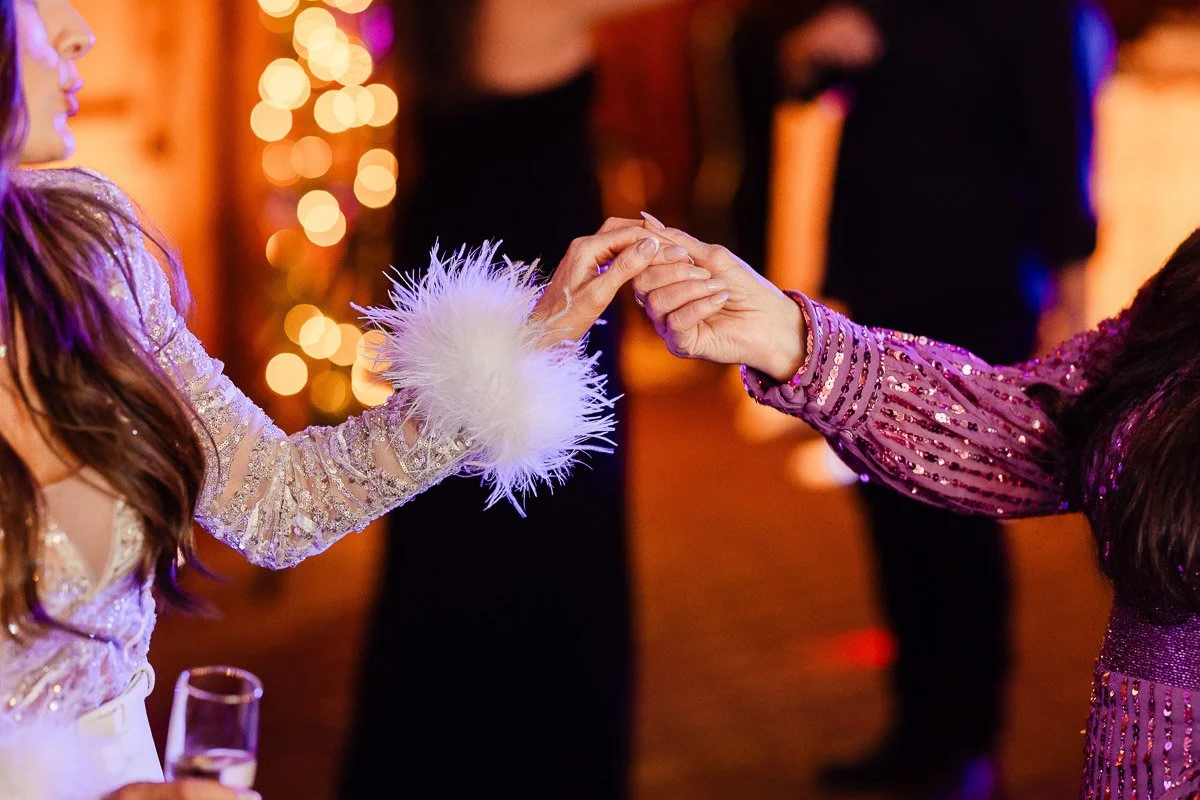 Two people touch hands at a festive event. One wears a sequined outfit with feather cuffs, the other is in a sparkly dress. Warm, joyful atmosphere.