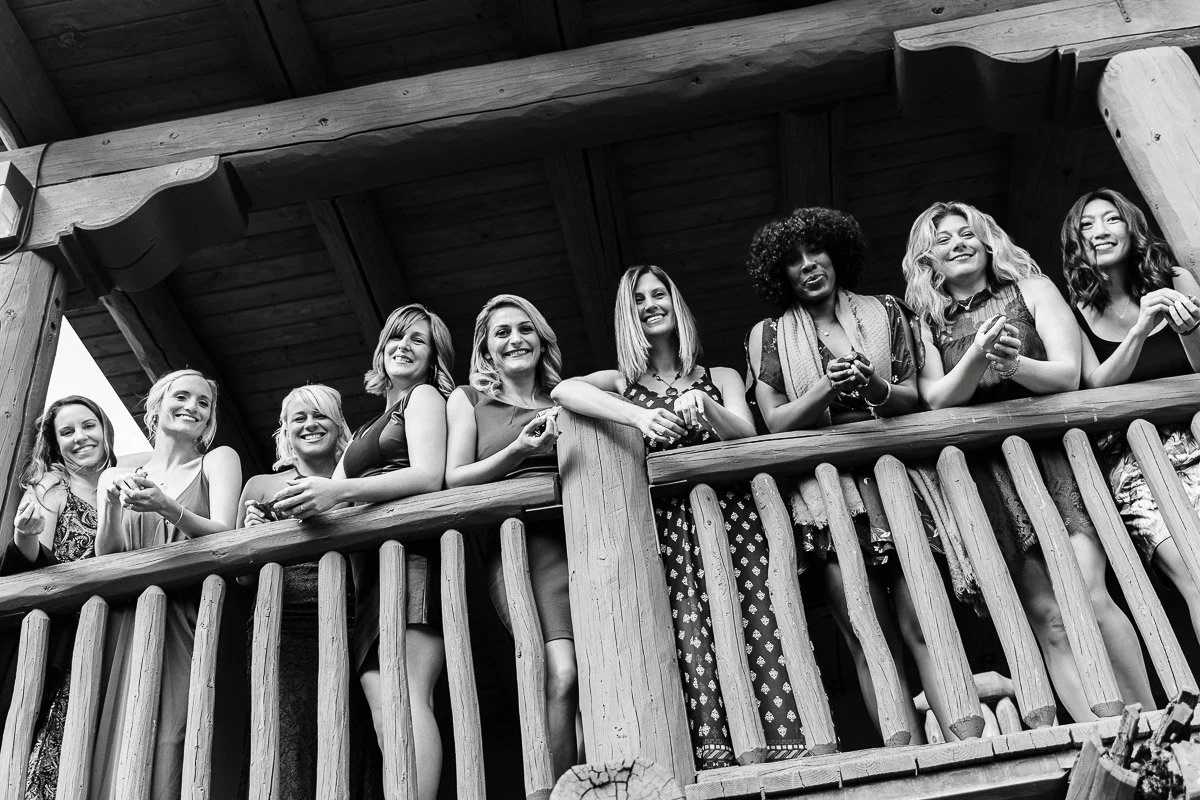 A black-and-white image of eight smiling women leaning on a rustic wooden railing, exuding joy and camaraderie. They stand close together on a balcony.