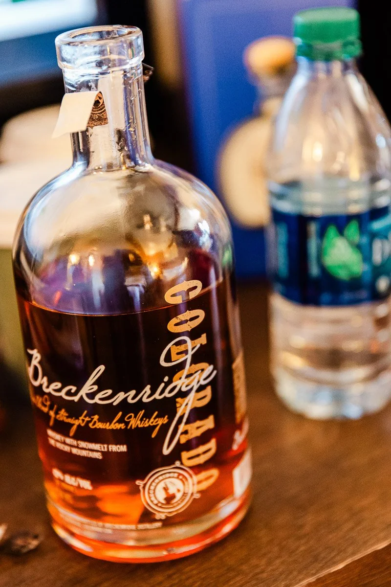 A bottle of Breckenridge bourbon whiskey with amber liquid sits on a wooden table beside a clear plastic water bottle, creating a relaxed and inviting mood.