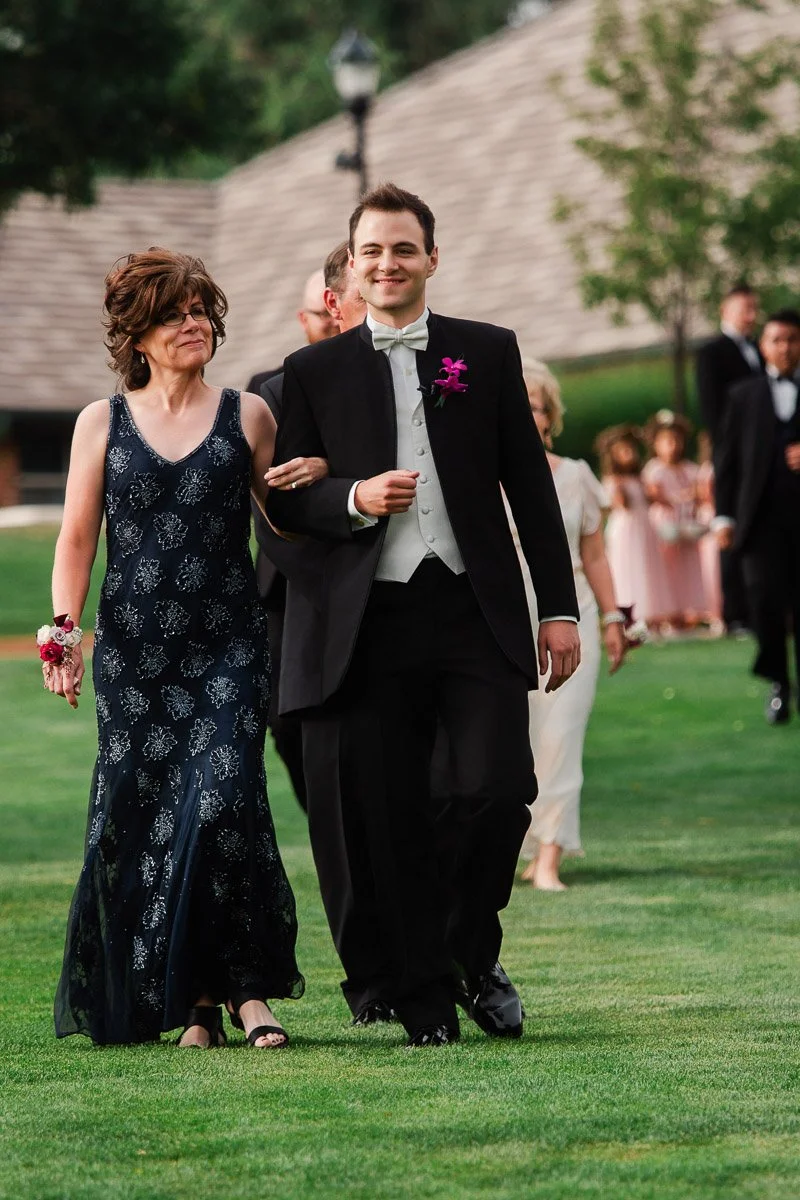 A man in a tuxedo and a woman in a navy gown walk arm in arm on a grassy lawn, surrounded by wedding guests dressed elegantly, exuding joy and celebration.