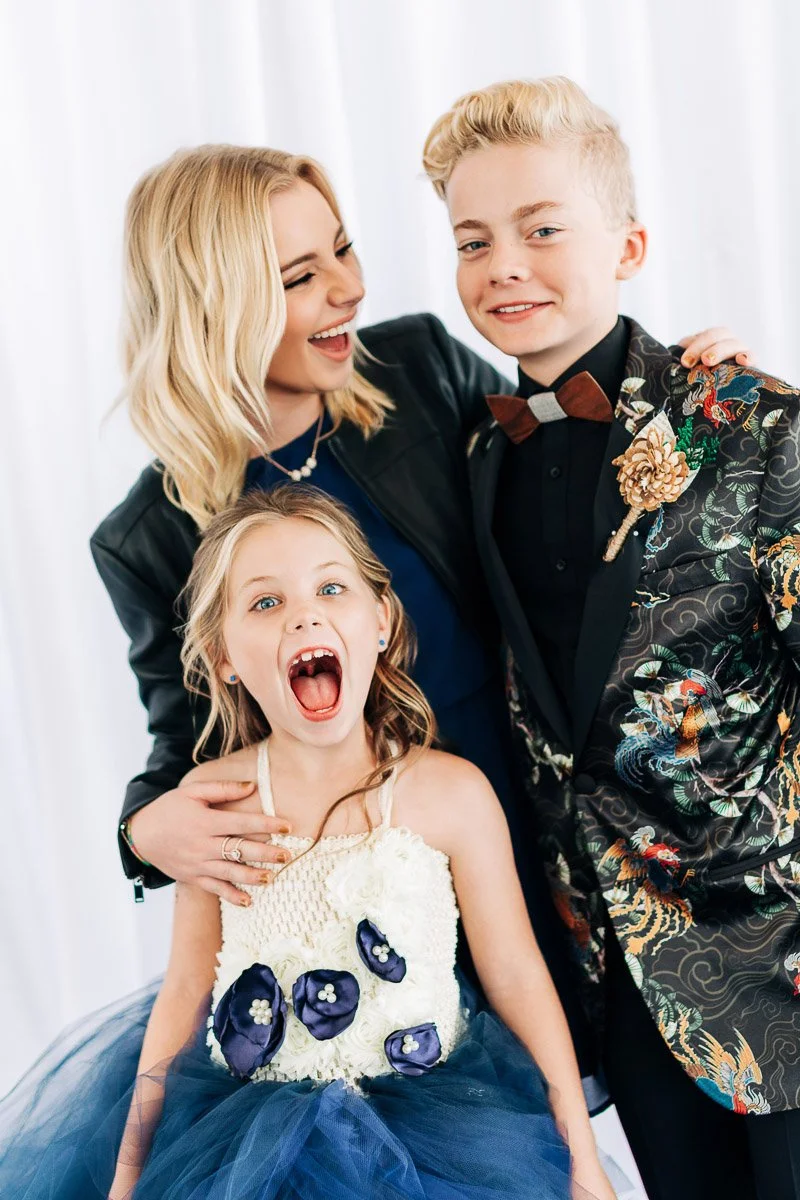 A smiling woman in a black jacket stands beside a young boy in a patterned suit with a bow tie. A girl in a blue and white dress playfully screams.