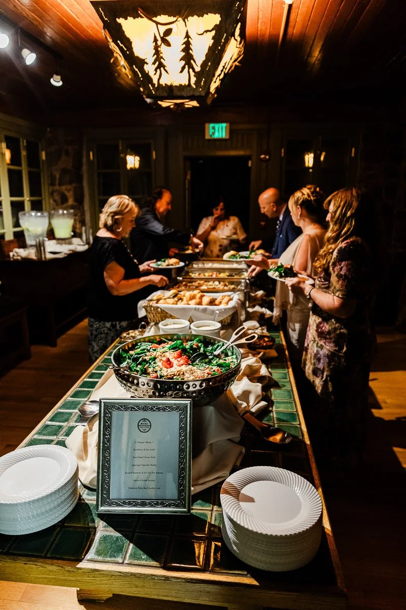 Buffet scene with people serving themselves under a warm, rustic light fixture. A table with salad, plates, and a menu adds to the cozy atmosphere.