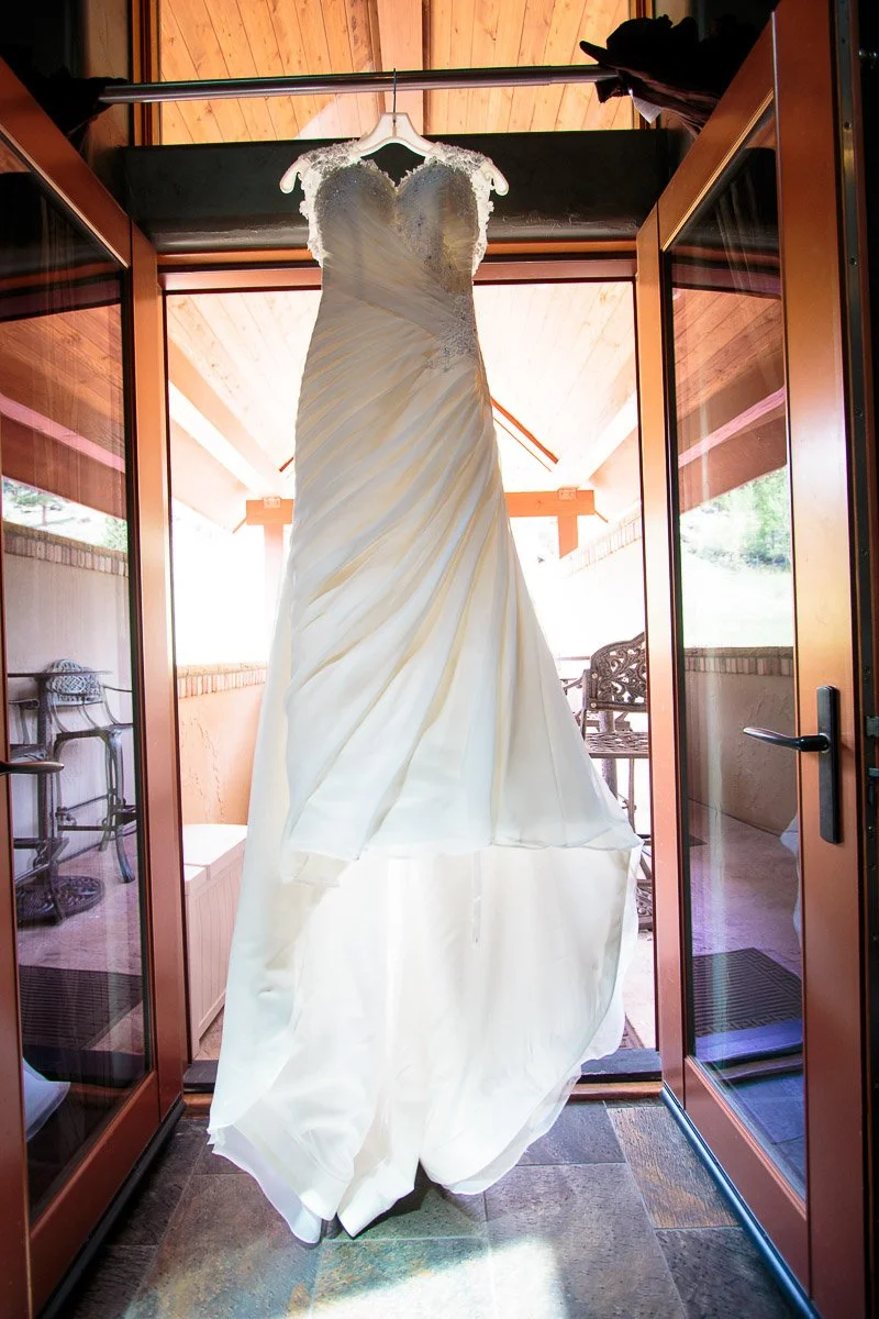 Elegant white wedding dress with lace straps and ruched bodice hanging in a sunlit doorway. Warm tones and serene outdoor view create a romantic ambiance.