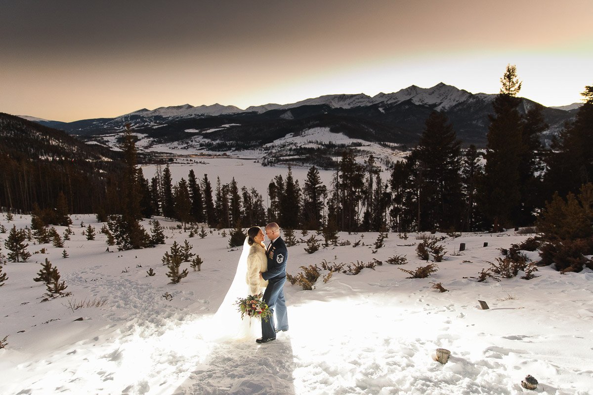 A couple embraces in a snowy mountain landscape at sunset, with shadows of tall trees and distant peaks. The scene is tranquil and romantic.