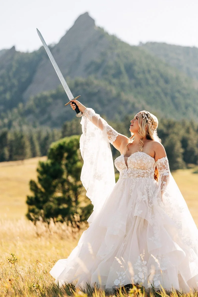 A woman in a flowing white gown holds a sword aloft in a meadow, with a mountain backdrop. The scene conveys empowerment and serenity captured by Boulder wedding photographer tomKphoto
