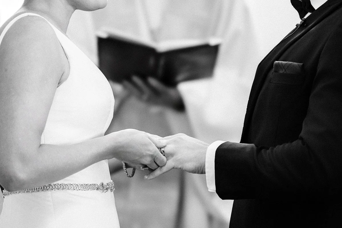 A bride and groom exchange rings during a wedding ceremony. They hold hands tenderly, with a priest in the background holding a book, creating a romantic and solemn atmosphere.