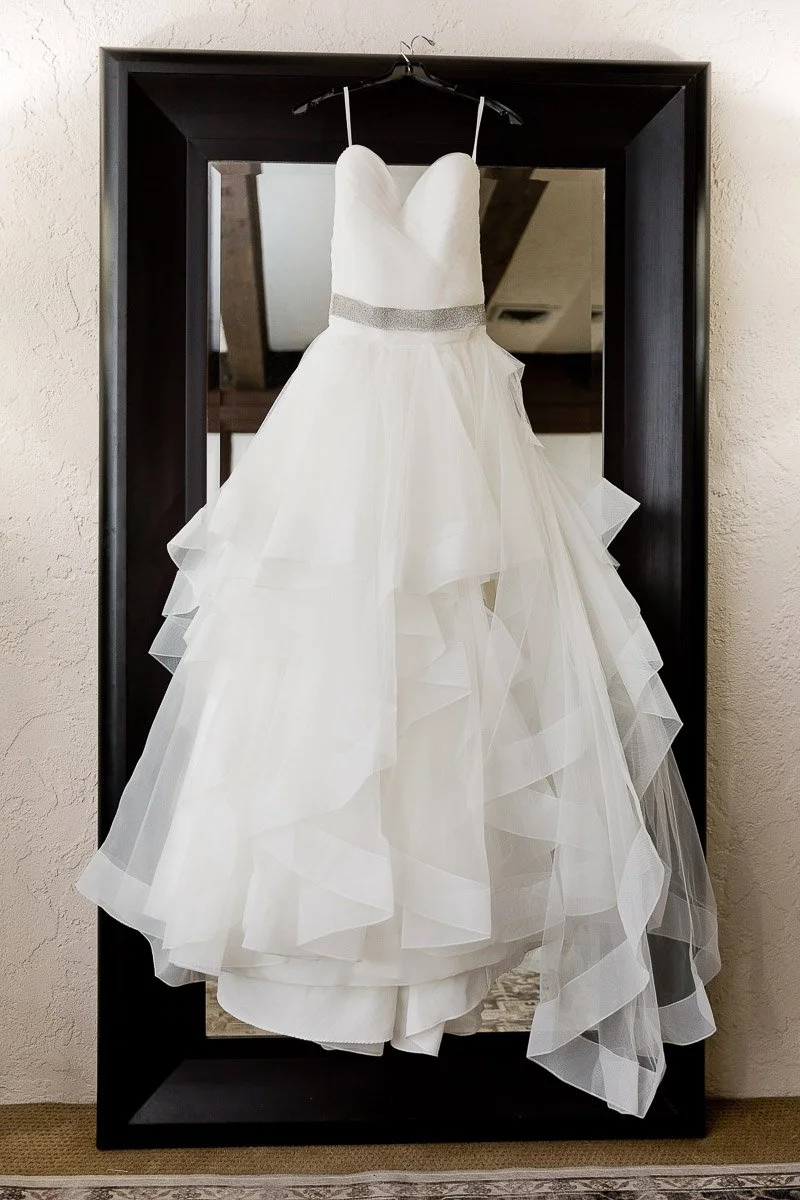 A strapless white wedding dress hangs elegantly on a mirror, featuring a layered tulle skirt and a silver waistband, set against a textured wall.